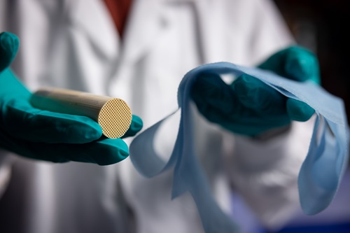 A close up of a personal in a white lab coat with blue gloves holding what looks like a cylinder in one hand and paper in the other.