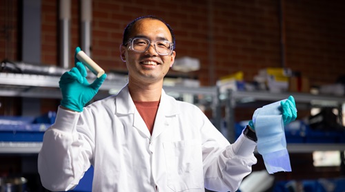 Dr Zhijian Wan in a white lab coat and blue gloves, holding a cylinder up in one hand and a piece of paper or material in the other, in a lab setting