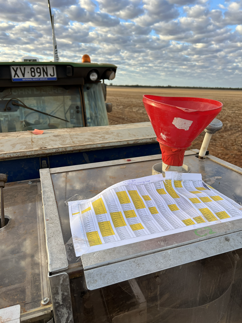 A printed trial plan, with some rows highlighted in yellow, is taped to the metal hopper of a plot seeder. A red funnel sits in the seed opening. A tractor is parked behind, and a bare, ploughed paddock stretches to the horizon under a sky of small, scattered clouds.