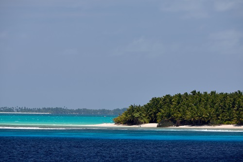 An island covered in palm trees surrounded by blue water of various colours.
