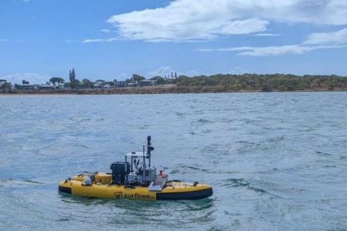 A yellow autonomous surface vessel equipped with scientific instruments travels across coastal waters near a low shoreline. The uncrewed vessel floats in the foreground, while trees, buildings and a partly cloudy blue sky form the background.