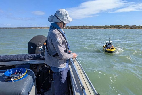 A person standing on the deck of a small research boat observes a yellow autonomous surface vessel travelling across calm coastal waters. A sandy shoreline and blue sky is visible in the background.