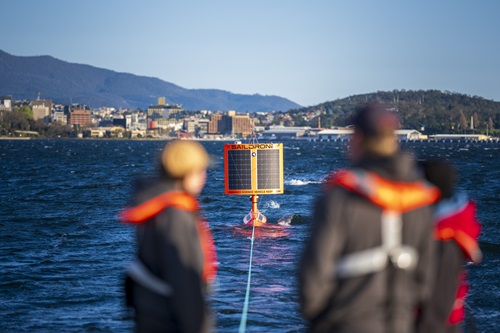 The USV on a tow line behind a boat leaves from Hobart to travel across the Southern Ocean. Three people blurred in the foreground on the boat wearing orange life jackets
