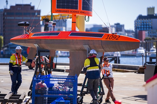 On a dock in Hobart with workers in hard hats and with high viz vests supporting the orange yacht-like USV as it is lifted from the dock.