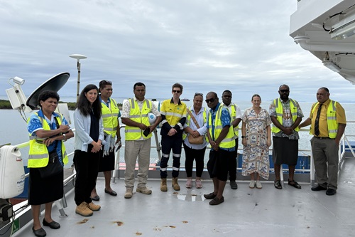 A group of people wearing yellow high-vis vests stand on the deck of a ship for a group photo.