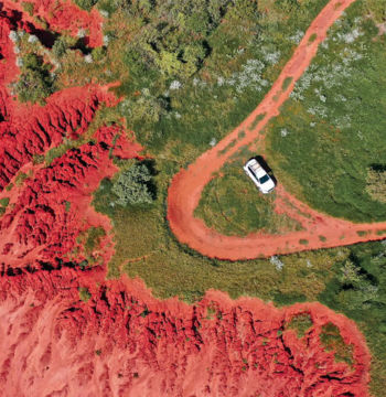 Overhead view of a white car driving on an orange dirt road with green open space leading to red hills