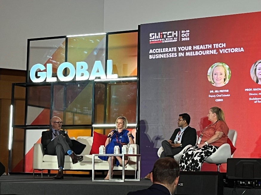 Four people with microphones sitting on couches on-stage at a conference. 