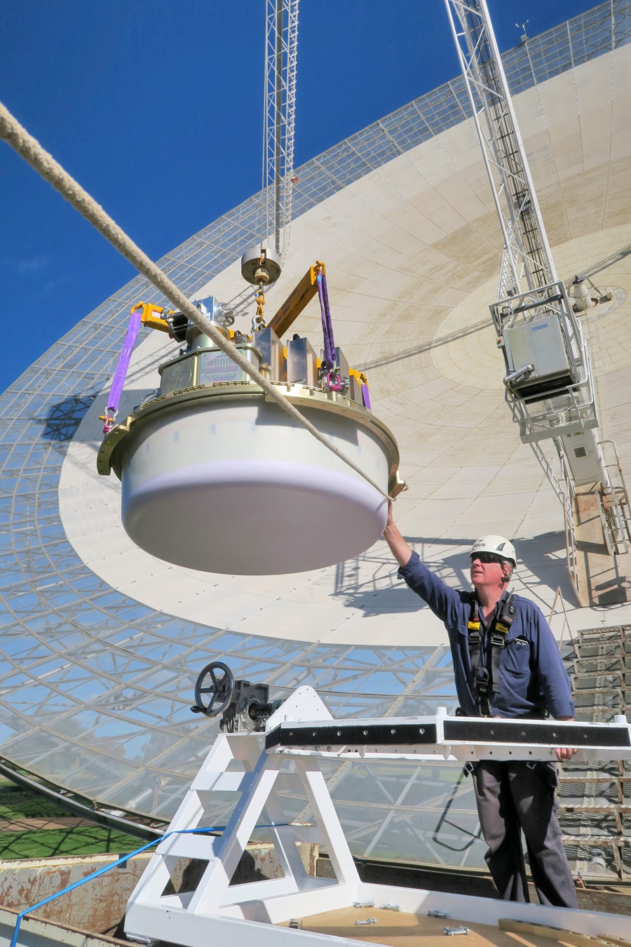 A man puts a hand to steady a large round object hanging in front of a large radio telescope.