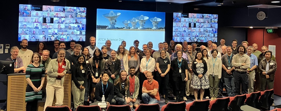 A large group of people gather in a lecture theatre with screens behind showing even more people and an image of a telescope.