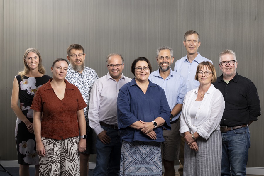 Nine people stand together, smiling towards the viewer