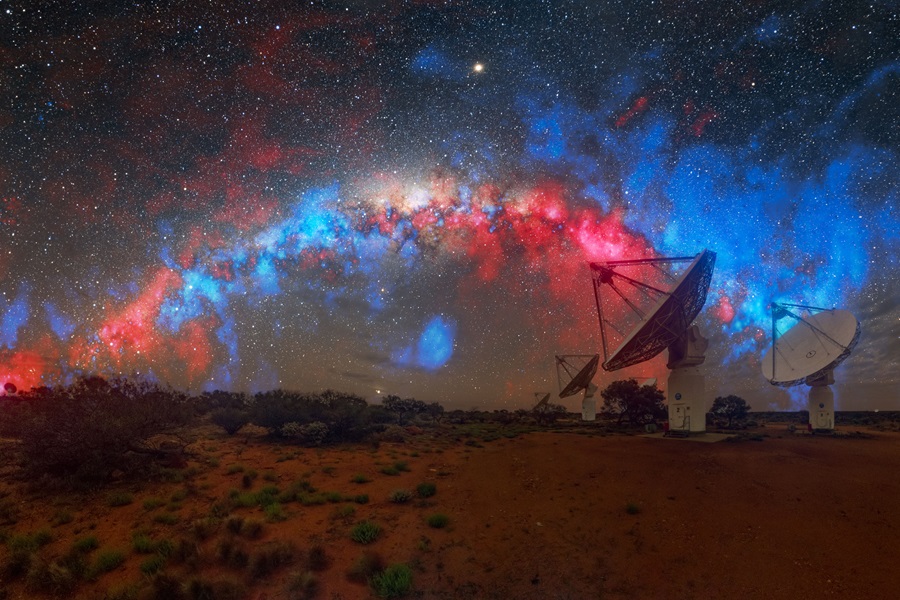 A nebulous cloud of red and blue arcs through a star-filled sky about telescope dishes