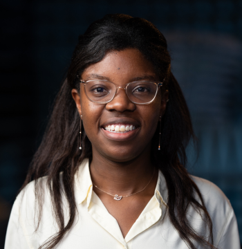 Natasha is a young black woman, she has glasses, wears a white shirt and is smiling at the camera