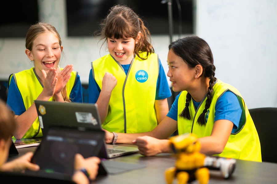 Three students wearing high-visibility vests gather around a laptop during a robotics activity, with a small robot visible on the table in the foreground.