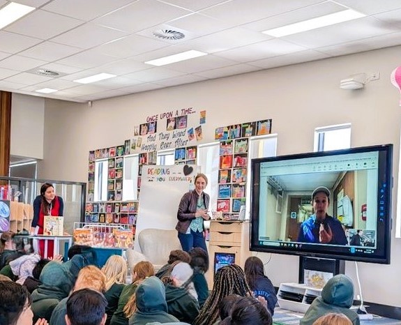 Students seated on a floor looking at a presenter, teacher and someone on a screen projected in front of them