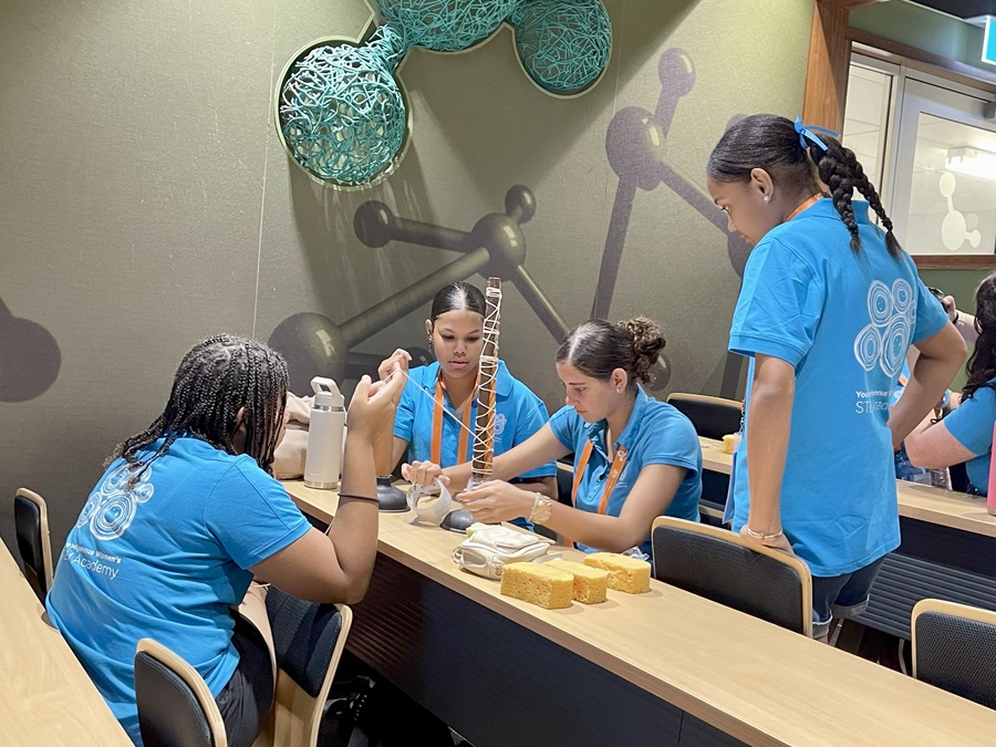Four students in blue STEM Academy shirts work together on a hands-on construction activity in a classroom. The students are seated and standing around a table, assembling a tall structure using string and other materials, with science-themed wall graphics visible in the background.