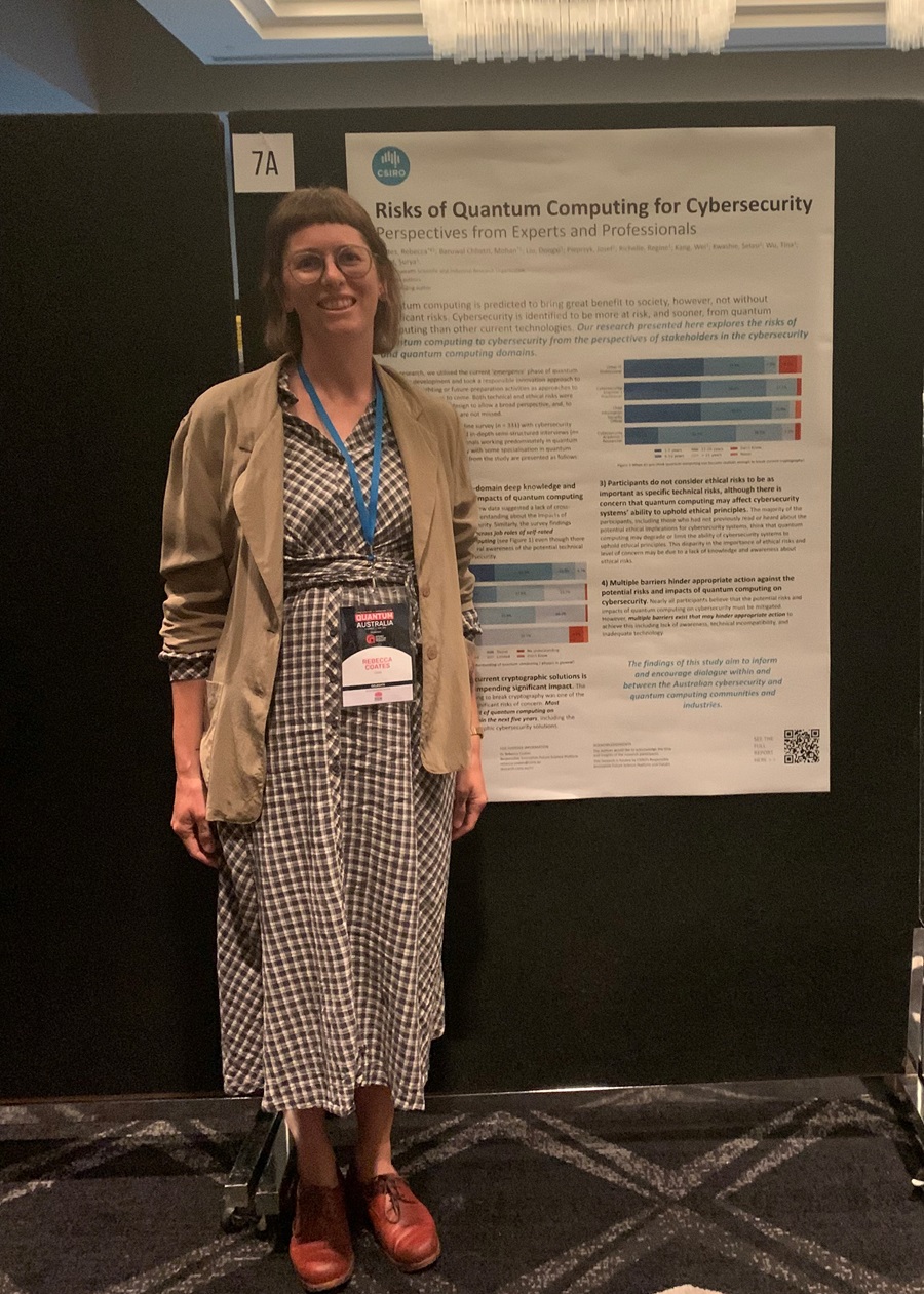 A female researcher wearing a long dress and blazer smiles standing next to a scientific poster. She is in a conference room and is wearing a lanyard.