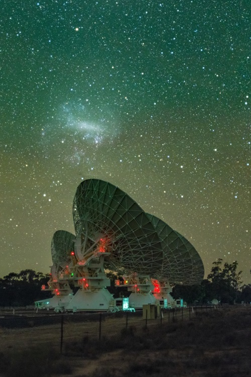 Five large radio telescope dishes face into a starry sky where a fuzzy cloud of a nearby galaxy is visible.