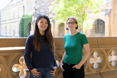 Two woman stand in front of a decorative stone balustrade. They are looking out at us and smiling. 