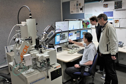 CSIRO researchers Mr Colin McRae (left), Dr Nick Wilson (middle), and Mr Aaron Torpy (right) look at data across six computer screens from the flagship electron probe microanalyser (EPMA), seen at left.