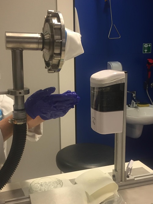 A pair of gloved hands apply hand sanitiser from a dispenser, directly beneath a piped fixture with a sealed face mask attached. 