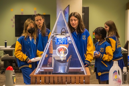 Students in school uniforms take turns putting recyclables into a bin with a robotic sorting arm