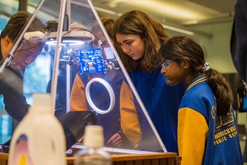 Students in school uniforms look through the glass top of a bin with a robotic sorting arm