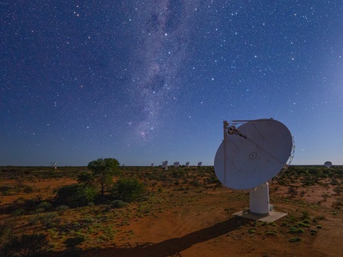 A dish antenna stands on desert soil beneath a starry sky. Other dish antennas are in the distance along the horizon