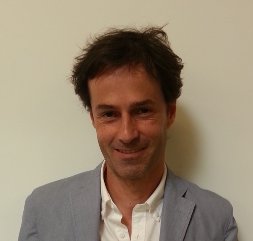 A man in a white shirt and blue jacket stands against a white wall