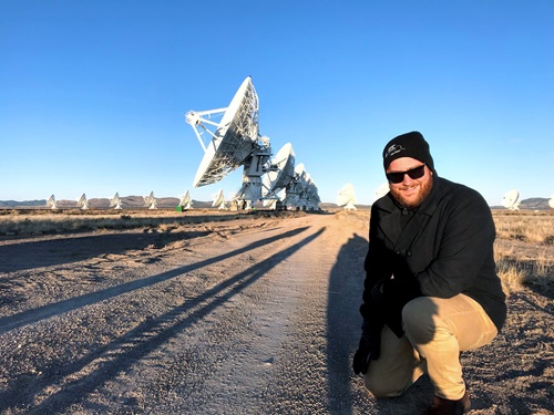 Man in beany and glasses poses in front of a radio telescope array