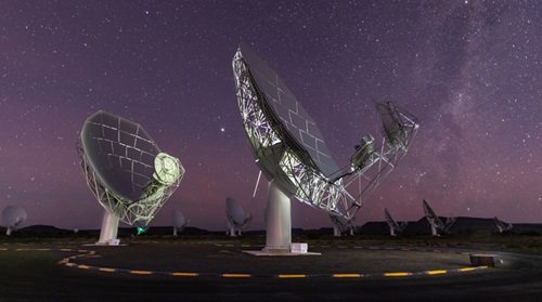 Two dish antenna in the foreground, stand underneath a starry sky. Other dishes are along the horizon behind them.