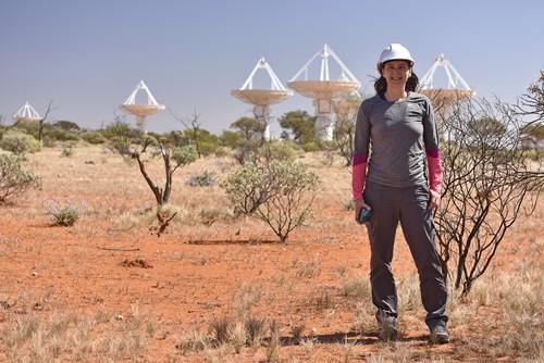 A woman stands in the desert. Behind her are dish antennas for a radio telescope