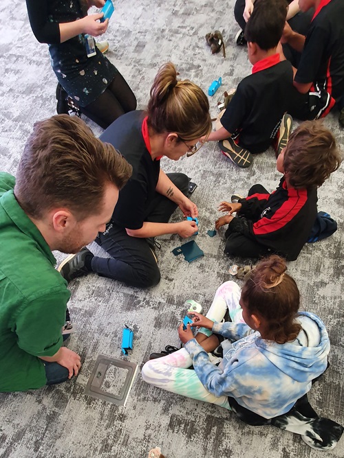 students in black and red uniforms sitting on carpet constructing bright blue cardstock into a small device to look through at lights