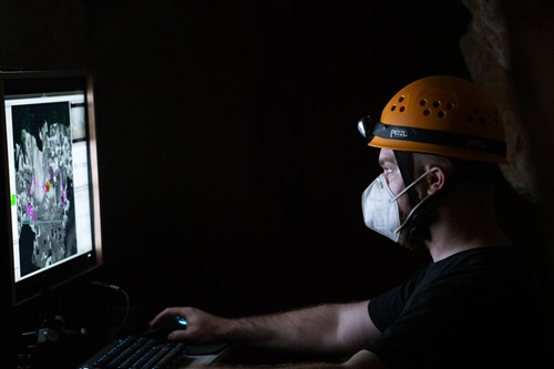 A scientist in a cave with a hard-hat and mask looking at a computer screen. 