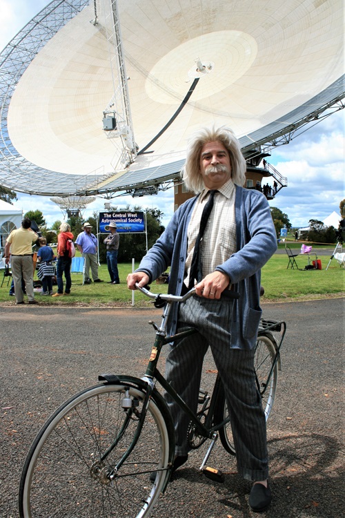 Science entertainer Patrick Helean dressed as Albert Einstein standing astride a bicycle in front of CSIRO's Parkes radio telescope, Murriyang.