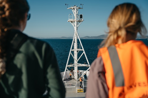 View from aboard a research vessel looking over the bow toward calm blue water and a distant coastline. Two trainees are visible in the foreground, with the ship’s mast centred in the frame and crew members working on deck below.