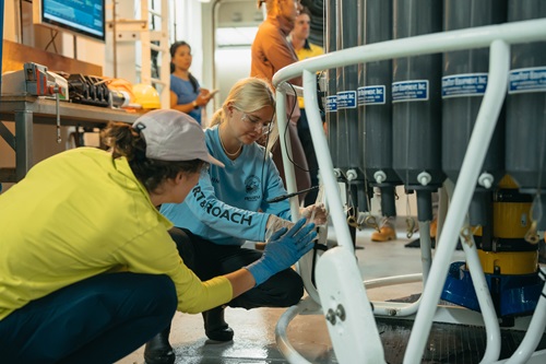 Two participants aboard a research vessel work with oceanographic sampling equipment inside a laboratory space. Wearing safety glasses and gloves, the participants adjust instruments mounted on a circular frame.
