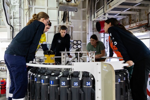 Five participants aboard a research vessel gather around a sampler in a shipboard laboratory.