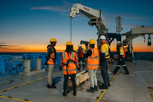 Several participants wearing high-visibility safety vests and hard hats stand on the aft deck of a research vessel during a training exercise. Shipboard cranes and scientific equipment are visible on deck, with the ocean and a colourful sunset on the horizon in the background.