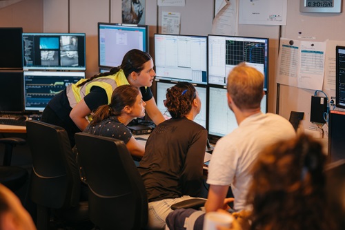 A group of participants aboard a research vessel observe data displays and monitoring screens in a shipboard operations room. Multiple computer monitors show scientific measurements, graphs and instrument readouts as participants review information together.