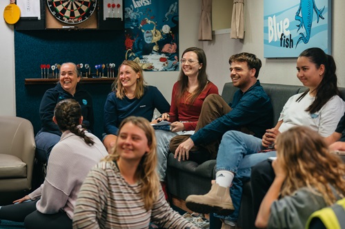 CAPSTAN participants sit together in a recreation room aboard RV Investigator. The group is gathered on couches and chairs in a relaxed setting featuring a dartboard and maritime-themed artwork, highlighting the social and community aspects of living and learning at sea during the training program.