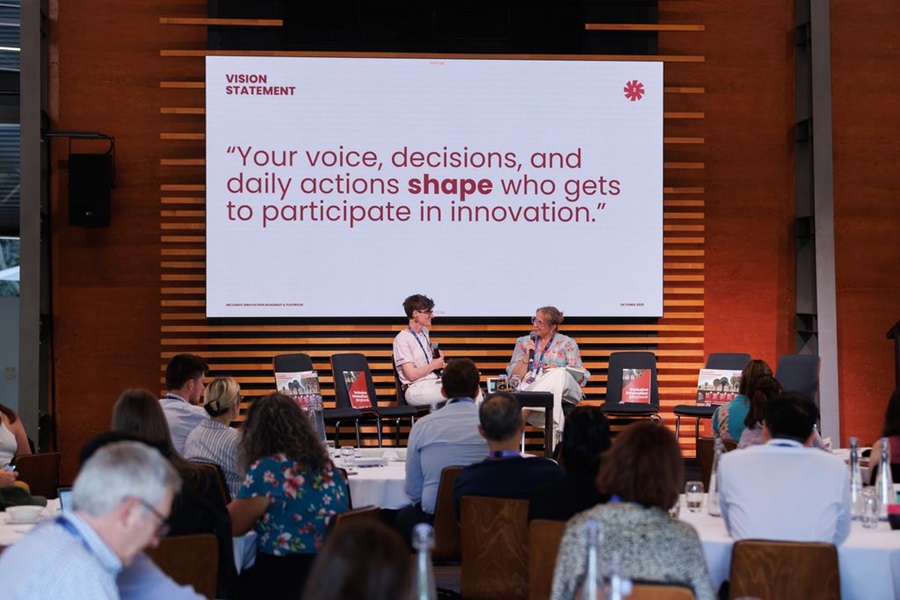 Two speakers sit on stage in front of a large screen displaying the text: “Your voice, decisions, and daily actions shape who gets to participate in innovation.” Audience members seated at round tables listen attentively during the Inclusive Innovation Roadmap & Playbook event hosted by CSIRO’s ON Innovation Program.