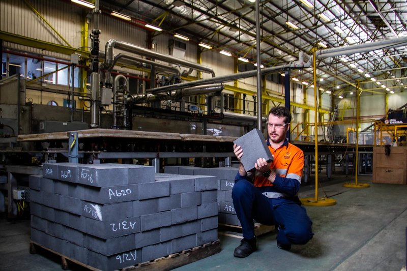 A worker inspects a thermal energy storage block in MGA Thermal's manufacturing facility, with stacks of storage blocks and industrial equipment in the background.