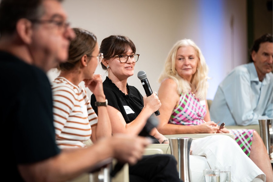 A woman sits on a panel with 4 other people. She is wearing back and reading glasses, with a microphone in her hand. She is holding it up as if speaking. The others are looking at her, listening. 