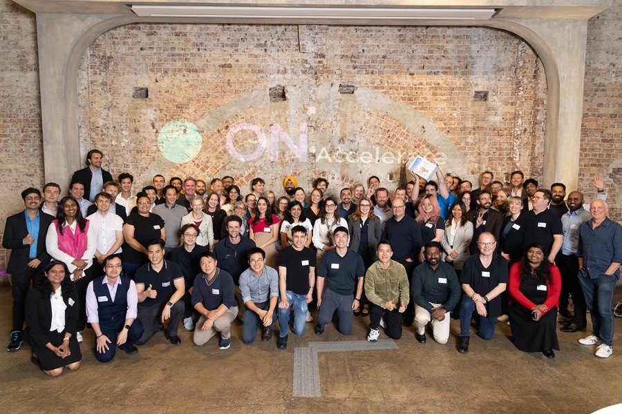 A large group of people posing together in front of a rustic brick wall with the words “ON Accelerate” projected on it. 