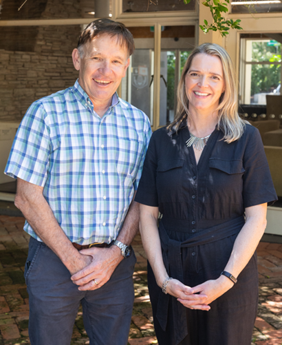 Two people standing outdoors on a brick patio in front of glass doors, wearing casual clothing.