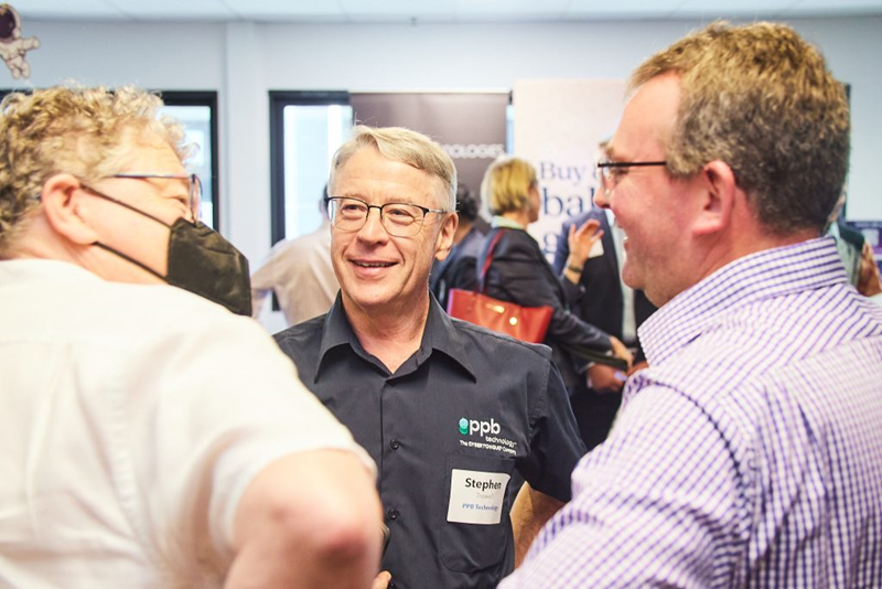 People engaged in conversation at a networking event, with a person wearing a dark shirt featuring the PPB Technology logo and a visible name badge. Background shows banners and other attendees.