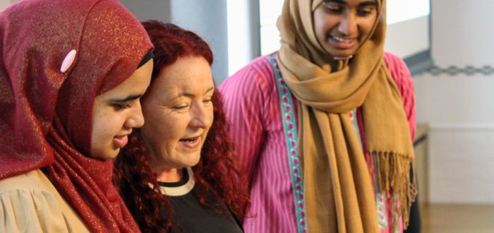 Three women stand together, looking down and talking together. The two women on both sides wear beautiful hijabs and clothing of red, pink and gold. The woman in the middle has brightly coloured red hair and a black top on and all look engaged in the discussion