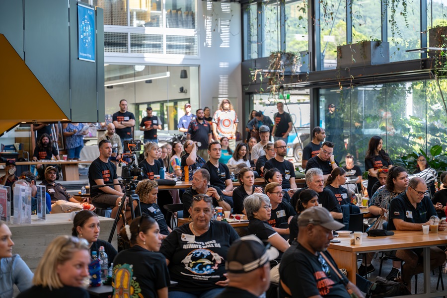 People gathered in a modern indoor venue with large glass windows and greenery outside, attending an event with tables, drinks, and cameras set up.