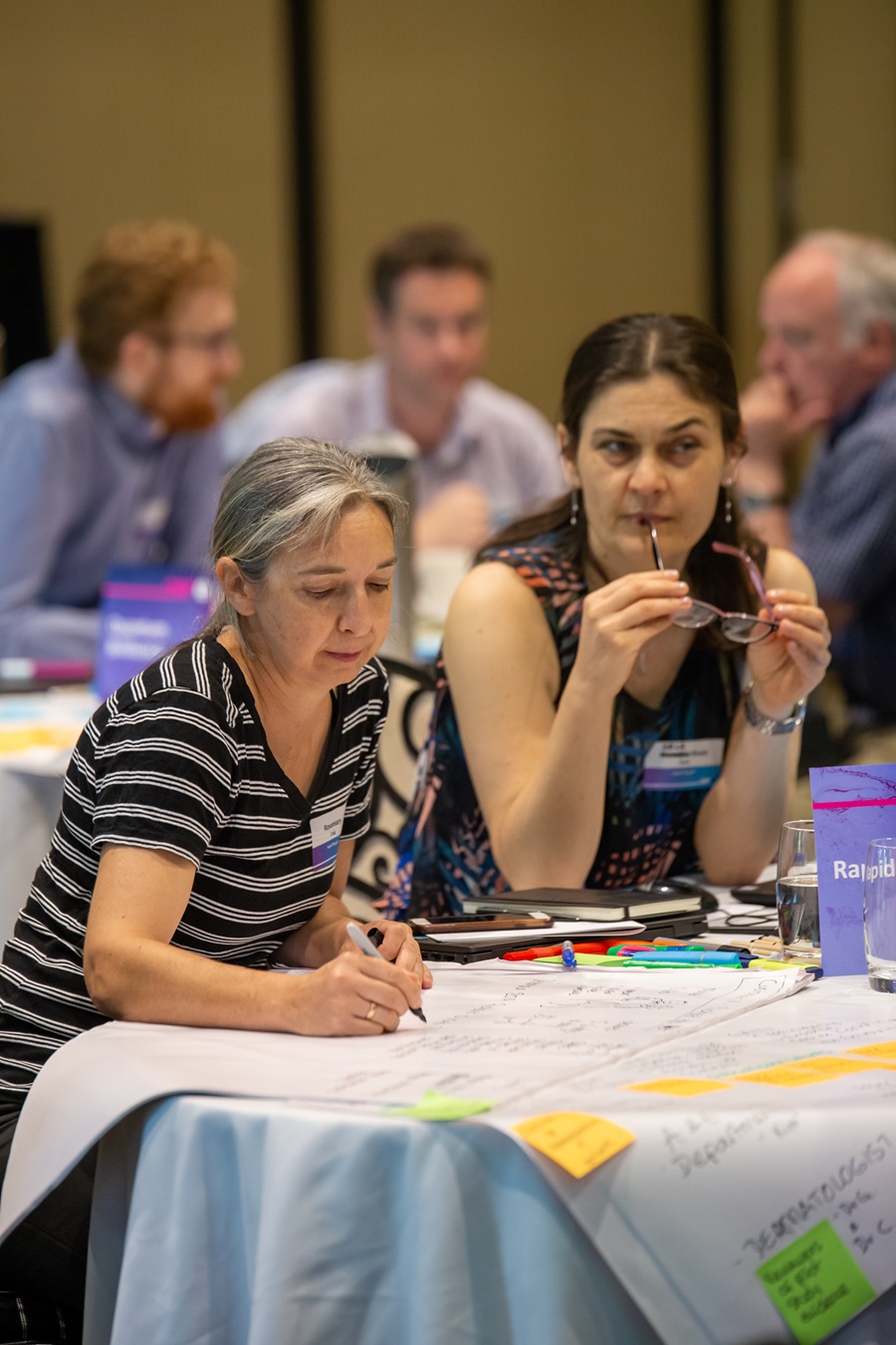 Two women sit at a table. They are writing on large pieces of paper on the table