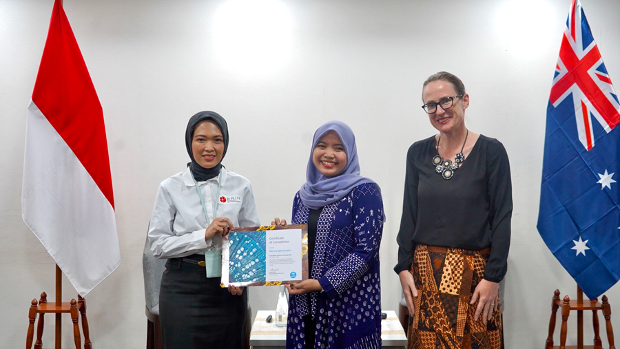Three women smiling at the camera while holding up a certificate, with the Indonesian and Australian flags are propped up on either side.